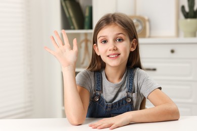 Photo of Happy little girl waving hello at white table indoors