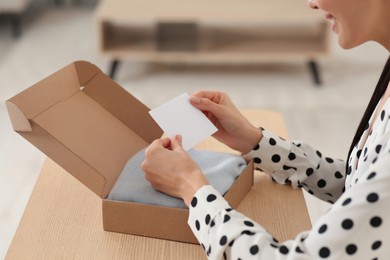 Happy woman holding greeting card near parcel with Christmas gift at table indoors, closeup