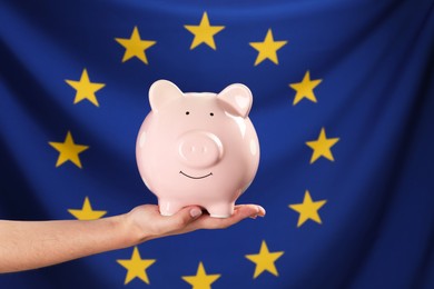 Photo of Woman holding pink piggy bank against flag of European Union, closeup