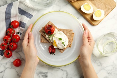 Woman holding fresh rye crispbreads with poached egg and grilled tomatoes at white marble table, top view