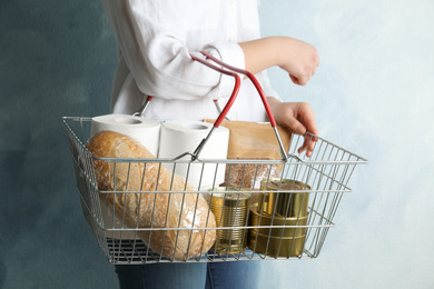 Photo of Woman holding shopping basket with products and toilet paper rolls on light blue background, closeup. Panic caused by virus