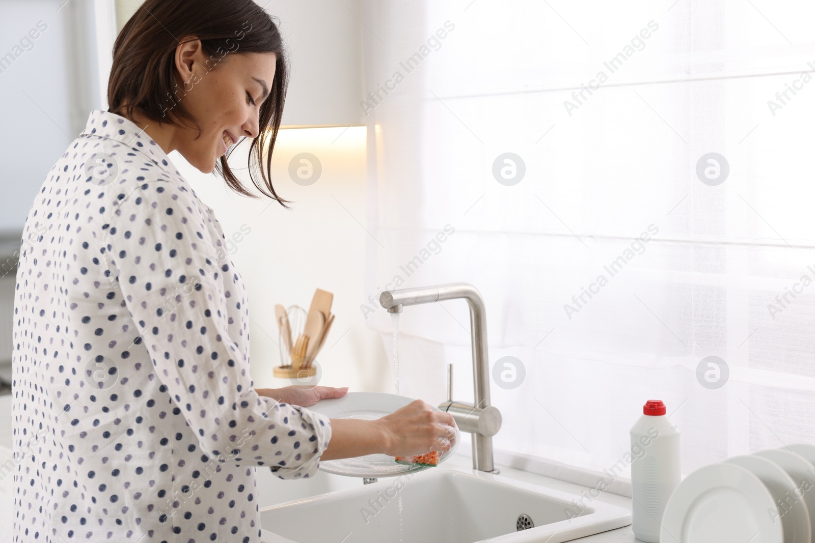 Photo of Happy young woman washing plate above sink in modern kitchen