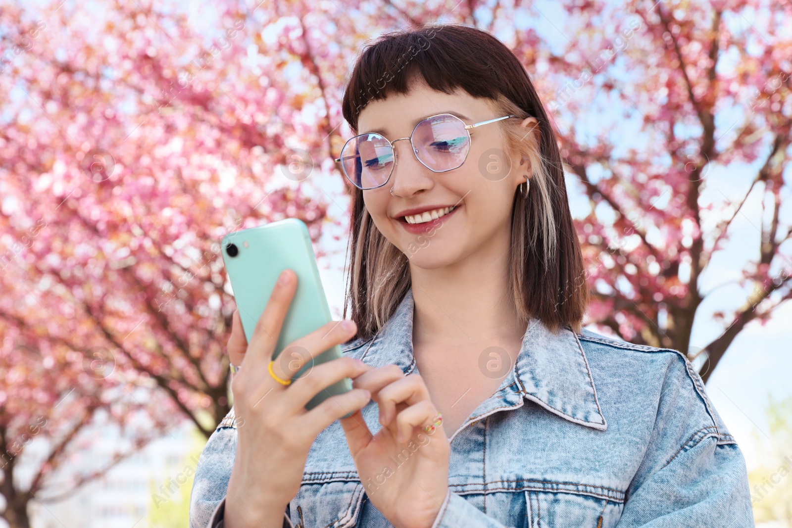 Photo of Beautiful young woman with phone near blossoming sakura tree in park