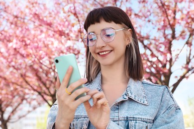 Beautiful young woman with phone near blossoming sakura tree in park