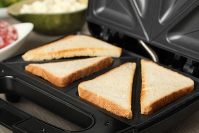 Photo of Modern sandwich maker with bread slices on wooden table, closeup