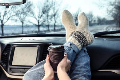 Photo of Young woman in warm socks holding her legs on car dashboard and drinking coffee. Cozy atmosphere