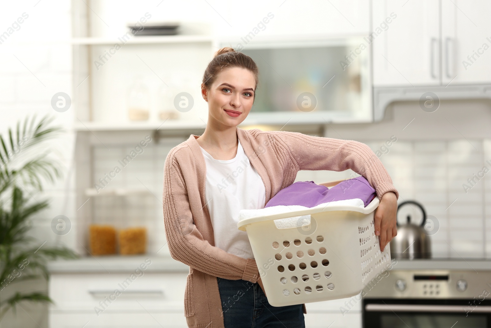 Photo of Woman holding basket with clean laundry in kitchen