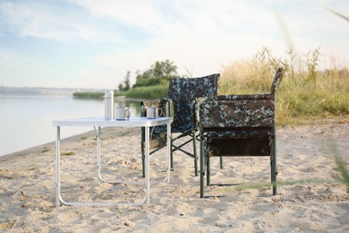 Photo of Camouflage fishing chairs and table with metal cups on sandy beach near river