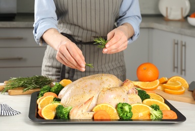 Photo of Woman adding rosemary to chicken with oranges and vegetables at table in kitchen, closeup