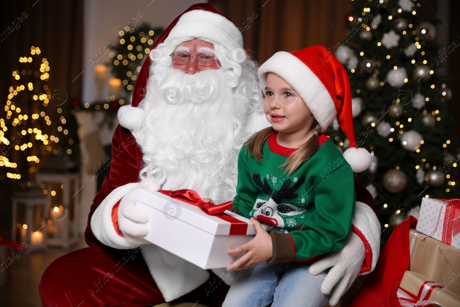 Photo of Santa Claus giving present to little girl in room decorated for Christmas