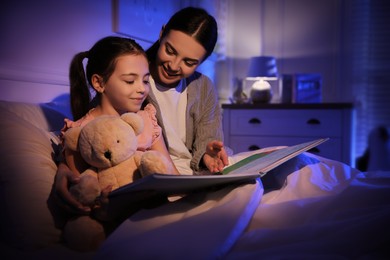 Mother with little daughter reading fairy tale in dark bedroom