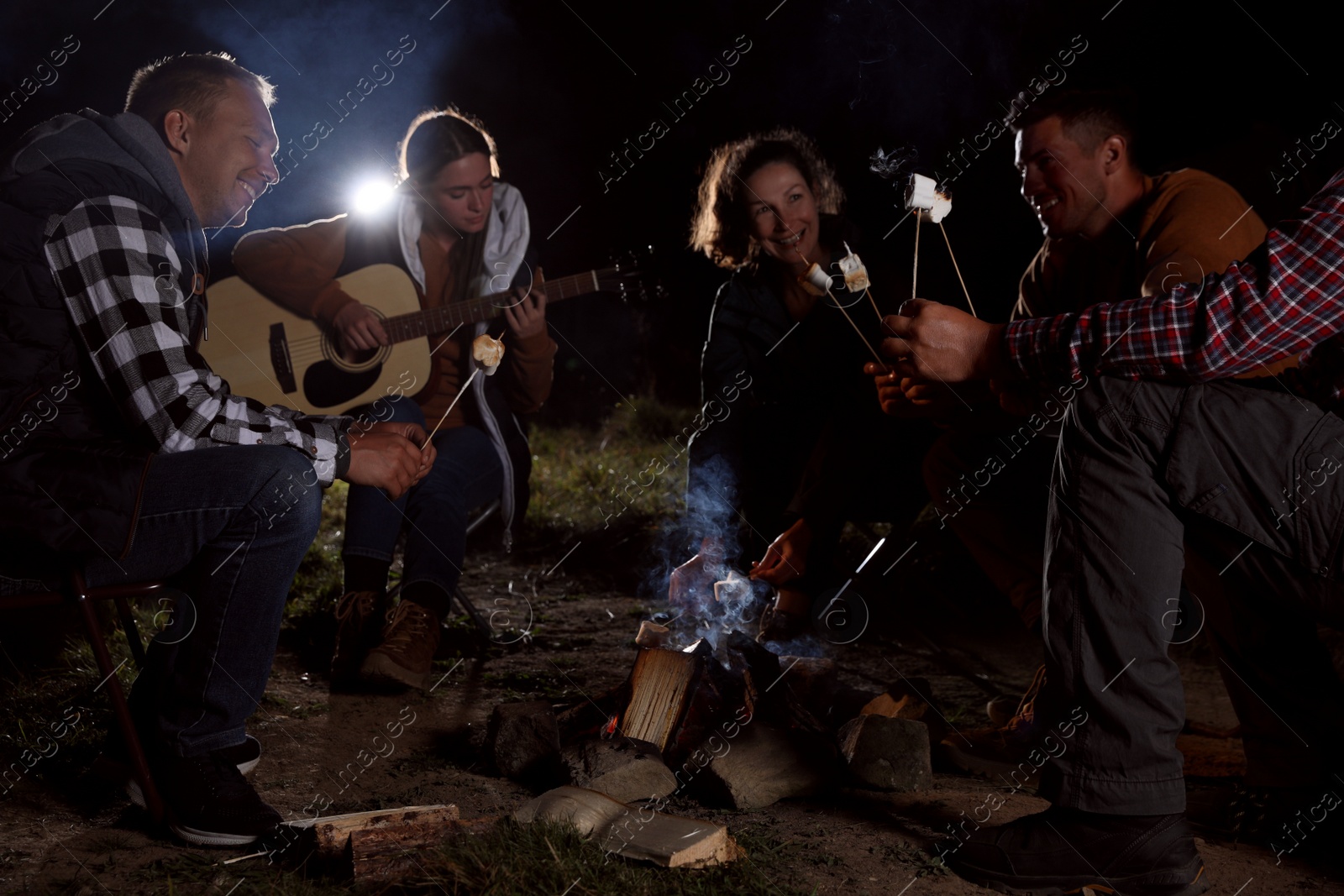 Photo of Group of friends roasting marshmallows on bonfire at camping site in evening