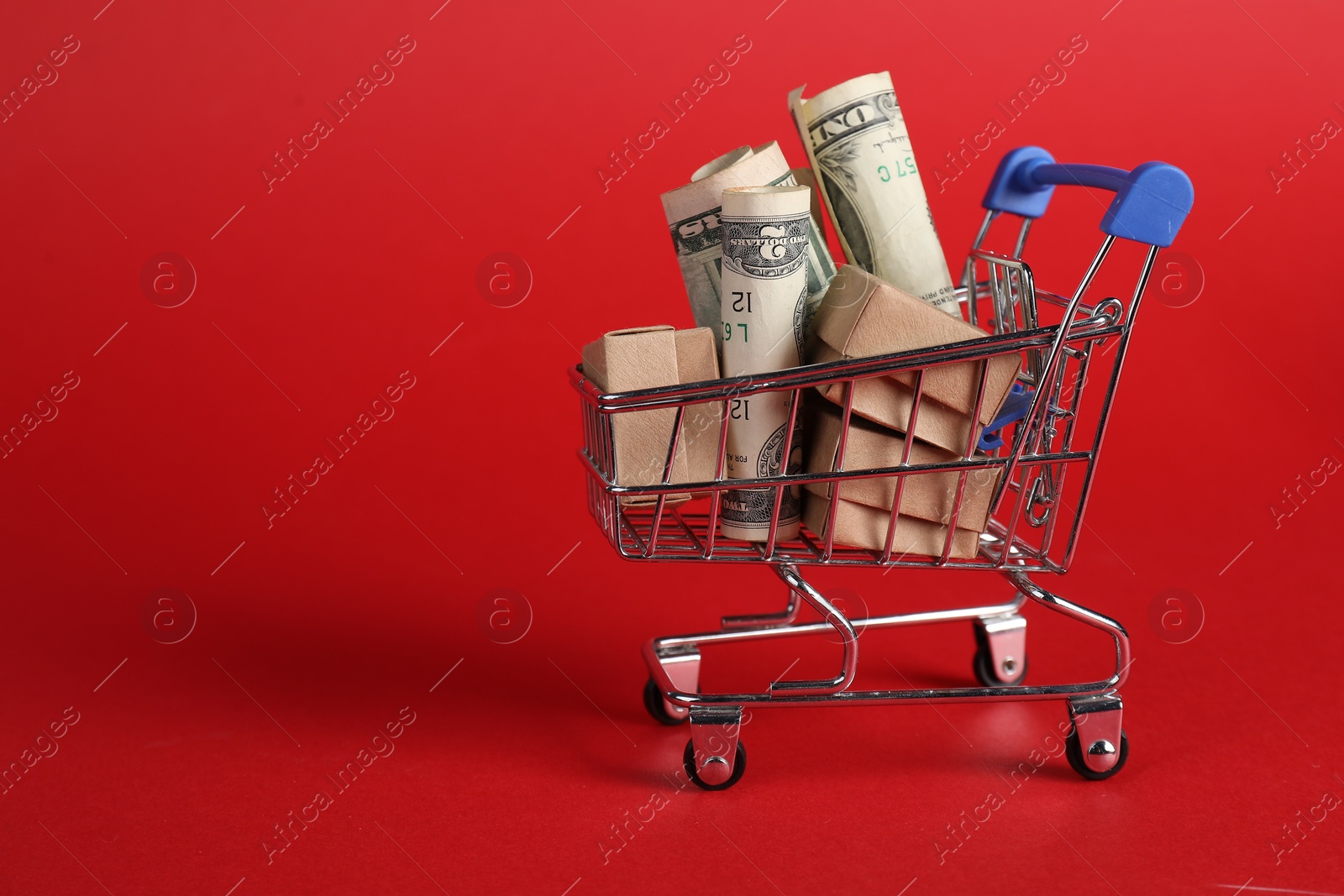 Photo of Small metal shopping cart with cardboard boxes and dollar banknotes on red background, space for text