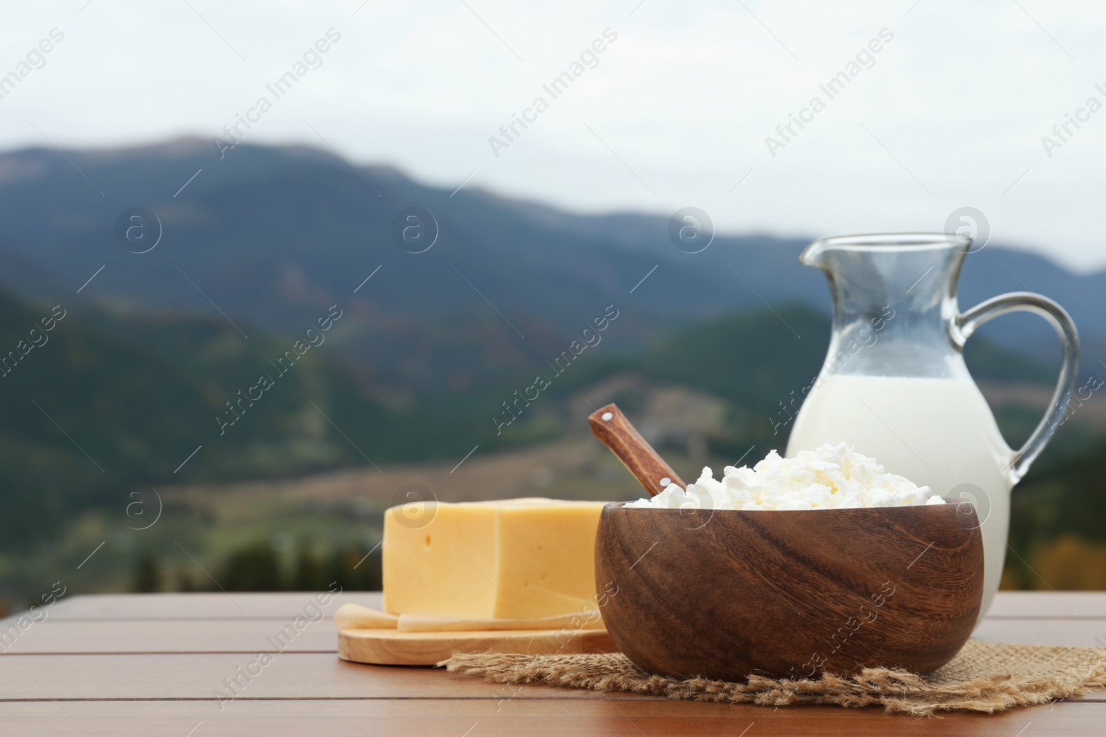 Photo of Tasty cottage cheese and other fresh dairy products on wooden table in mountains. Space for text