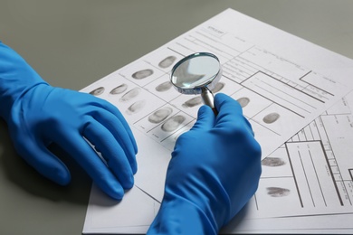 Photo of Criminalist exploring fingerprints with magnifying glass at table, closeup