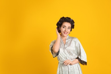 Photo of Happy young woman in silk bathrobe with hair curlers on orange background, space for text