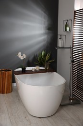 Stylish bathroom interior with ceramic tub, candles and care products on wooden bath tray