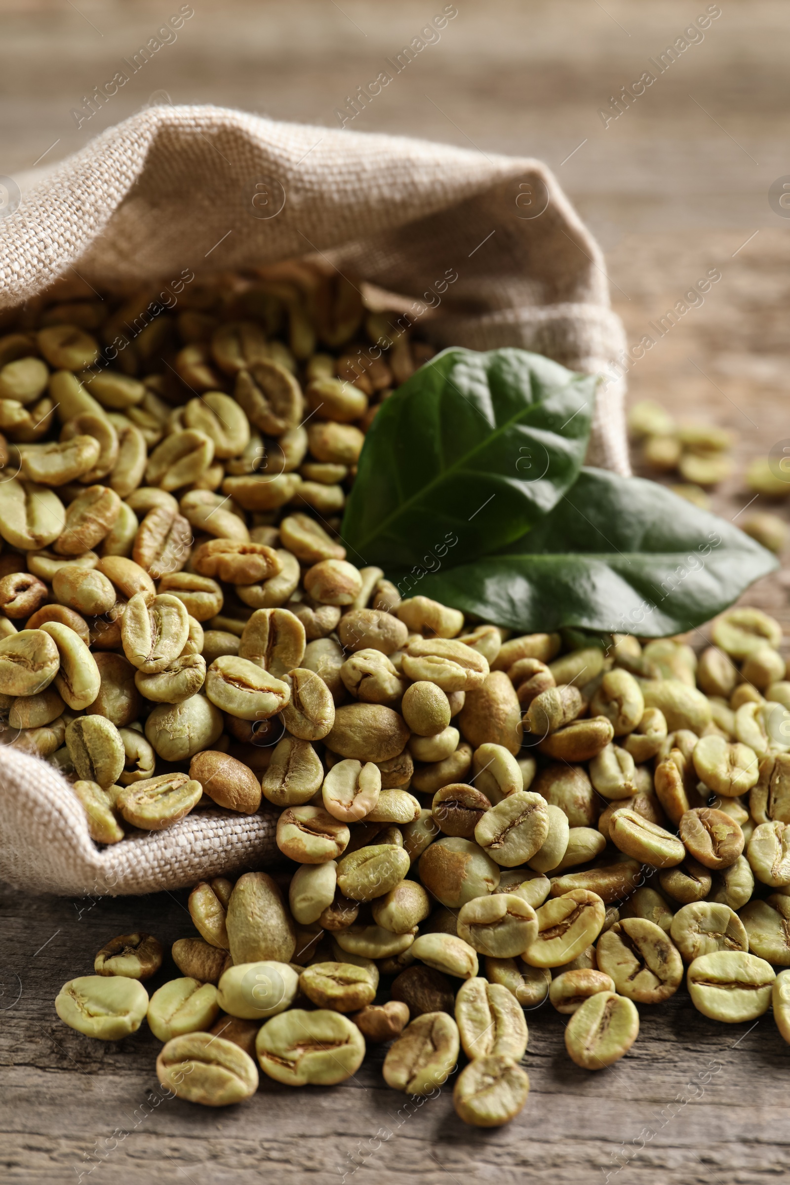 Photo of Overturned sackcloth bag with green coffee beans and leaves on wooden table, closeup