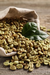 Photo of Overturned sackcloth bag with green coffee beans and leaves on wooden table, closeup