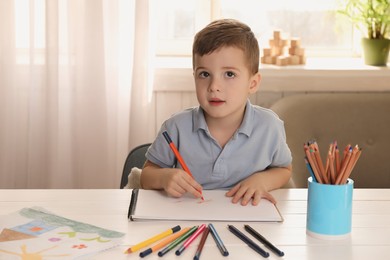 Photo of Cute little boy drawing with pencil at white wooden table in room. Child`s art