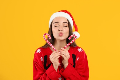 Photo of Young woman in Christmas sweater and Santa hat holding candy canes on yellow background