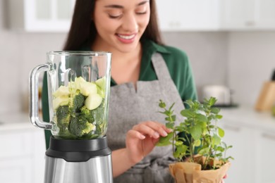 Photo of Woman holding potted herb indoors, focus on blender with ingredients for smoothie