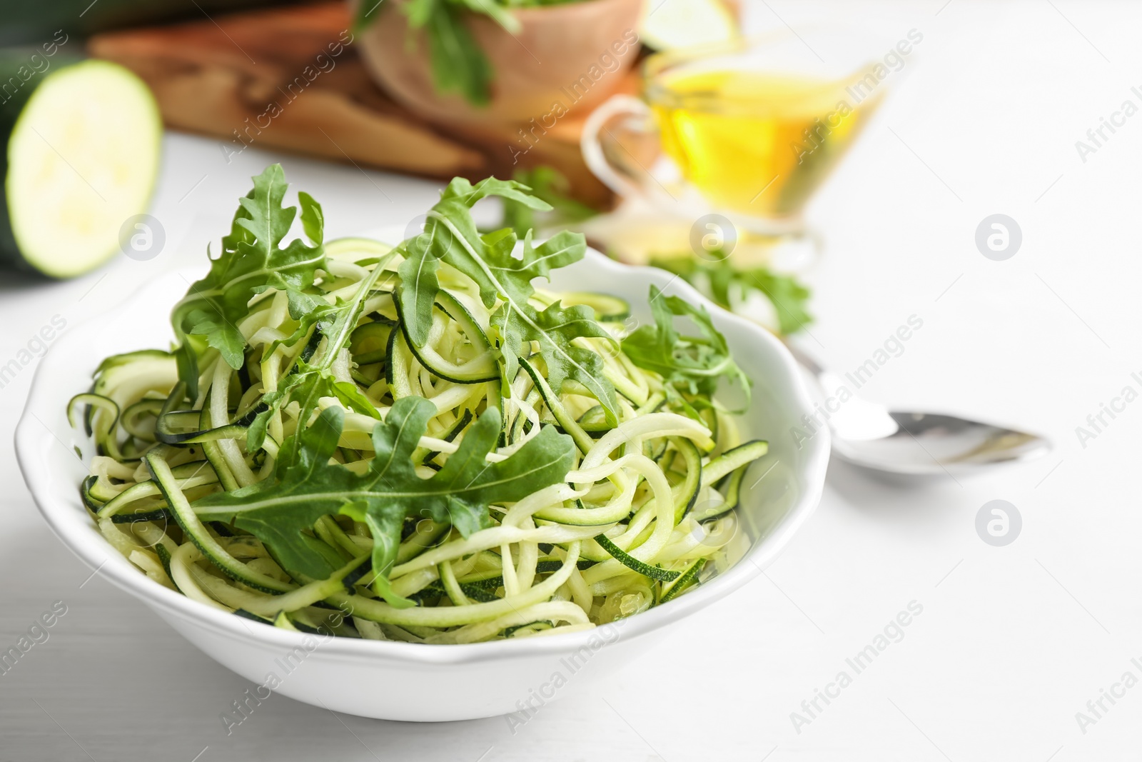 Photo of Delicious zucchini pasta with arugula in bowl on white wooden table, closeup