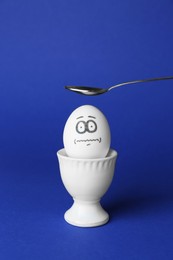 Photo of Spoon over egg with drawn frightened face among others in cup on blue background