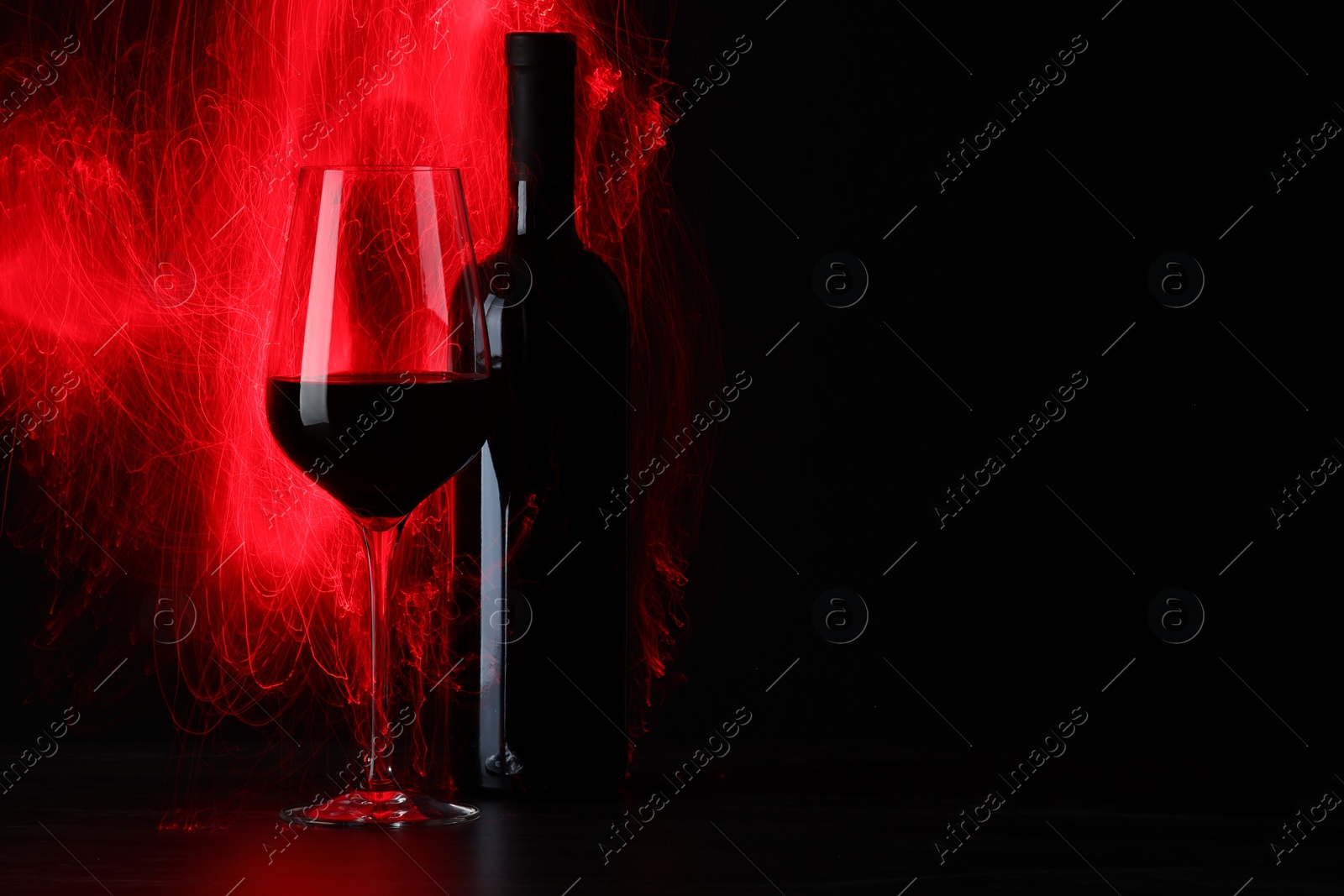 Photo of Tasty wine in glass and bottle in red lights on black background, space for text