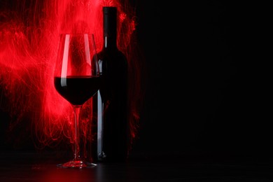 Tasty wine in glass and bottle in red lights on black background, space for text
