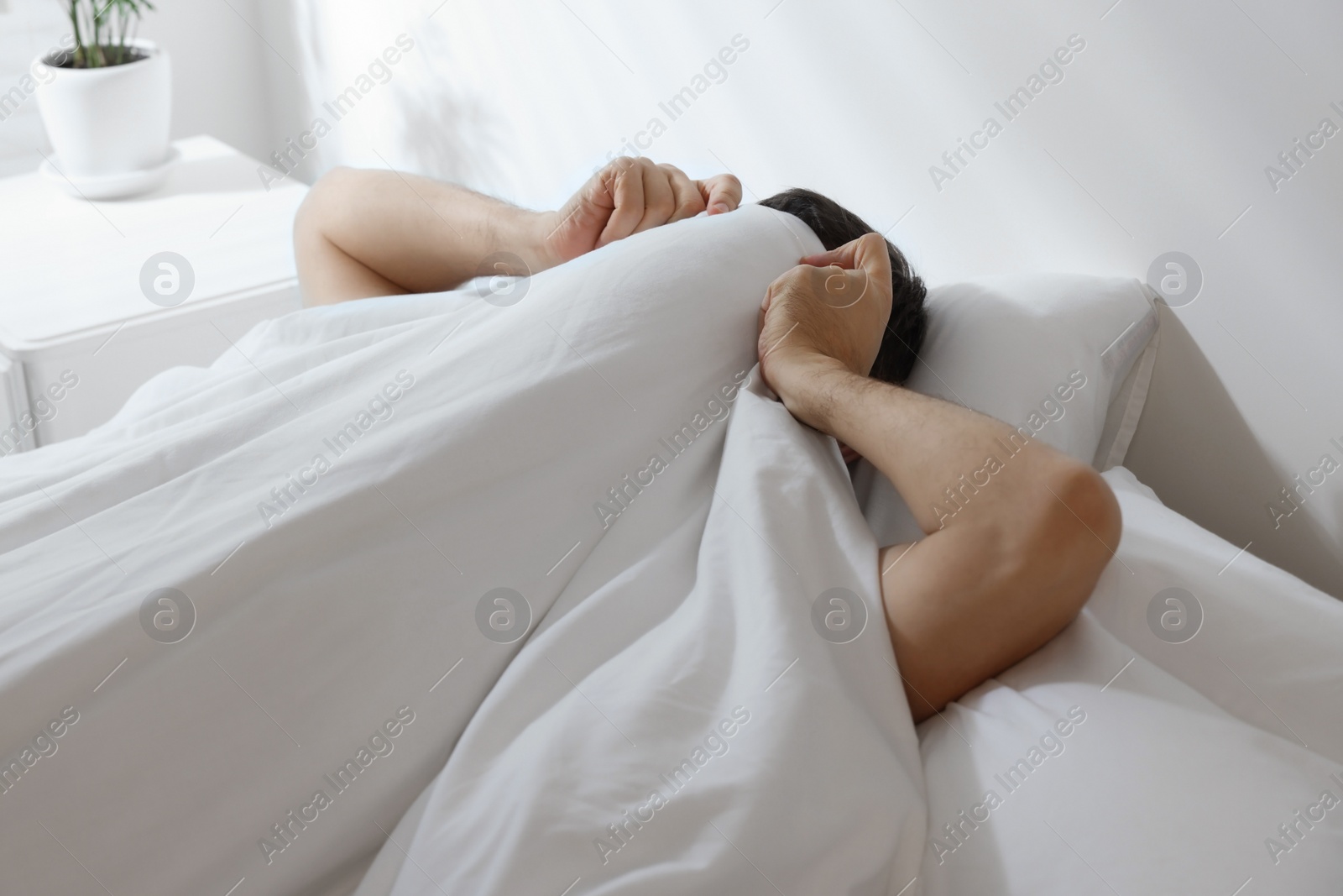 Photo of Sleepy man covering his face with blanket in bed at home