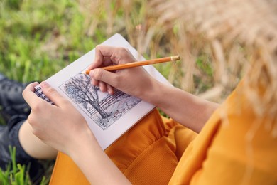 Photo of Young woman drawing with pencil in notepad outdoors, closeup