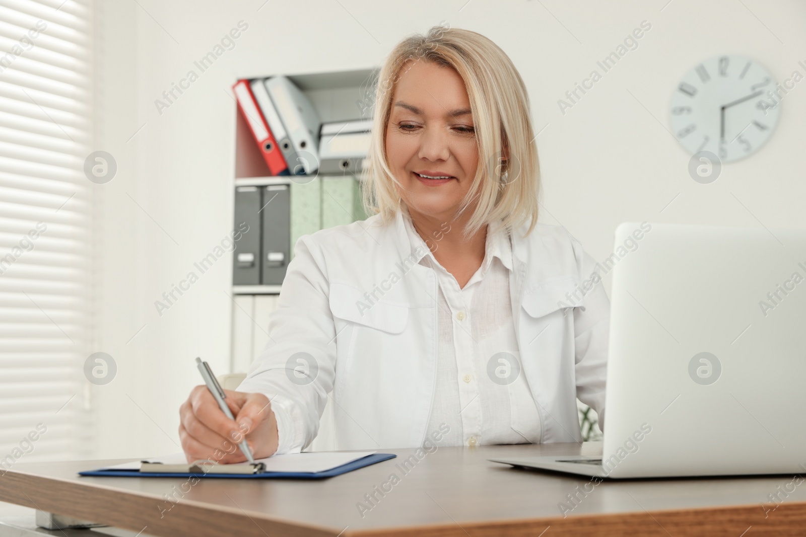 Photo of Doctor with laptop and clipboard writing at wooden table in clinic