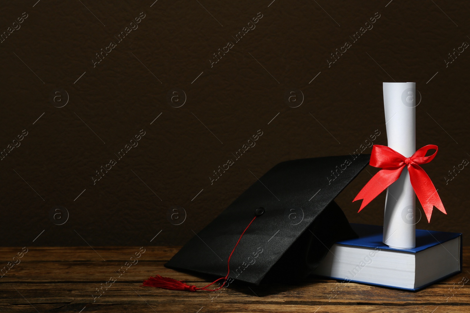 Photo of Graduation hat, book and diploma on wooden table against brown background, space for text