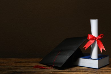 Photo of Graduation hat, book and diploma on wooden table against brown background, space for text