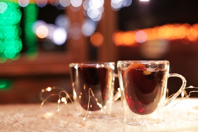 Glass cups of mulled wine and garland on table covered with snow outdoors. Space for text