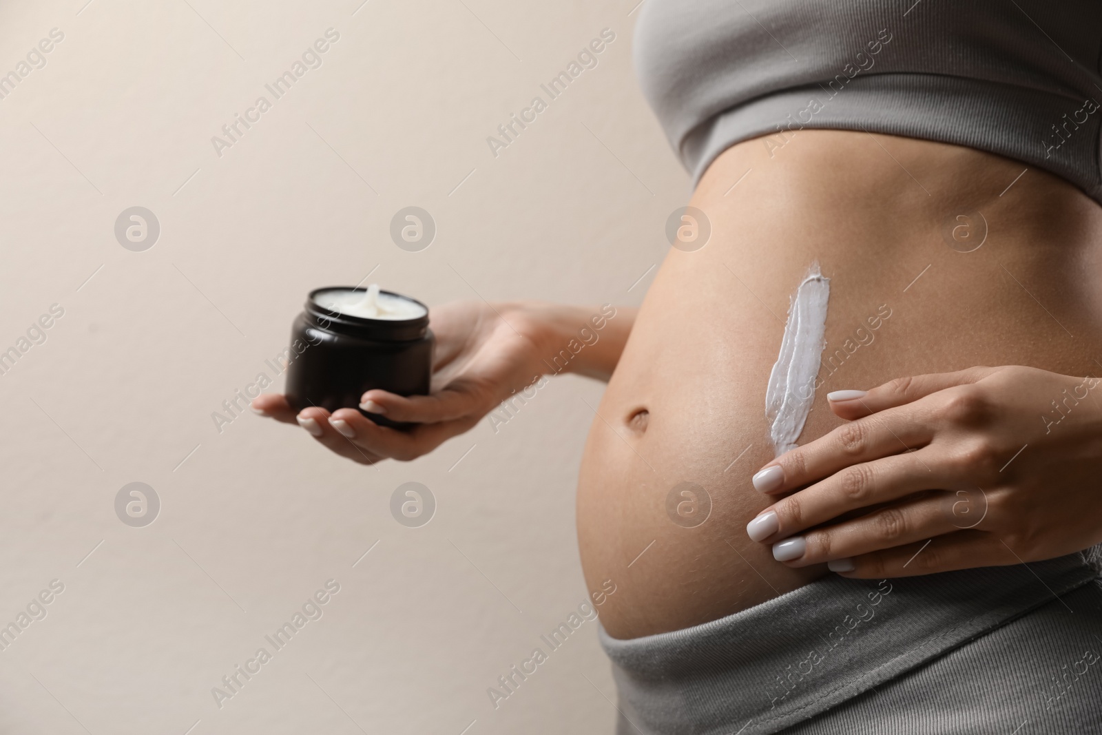 Photo of Pregnant woman applying cosmetic product on belly against beige background, closeup