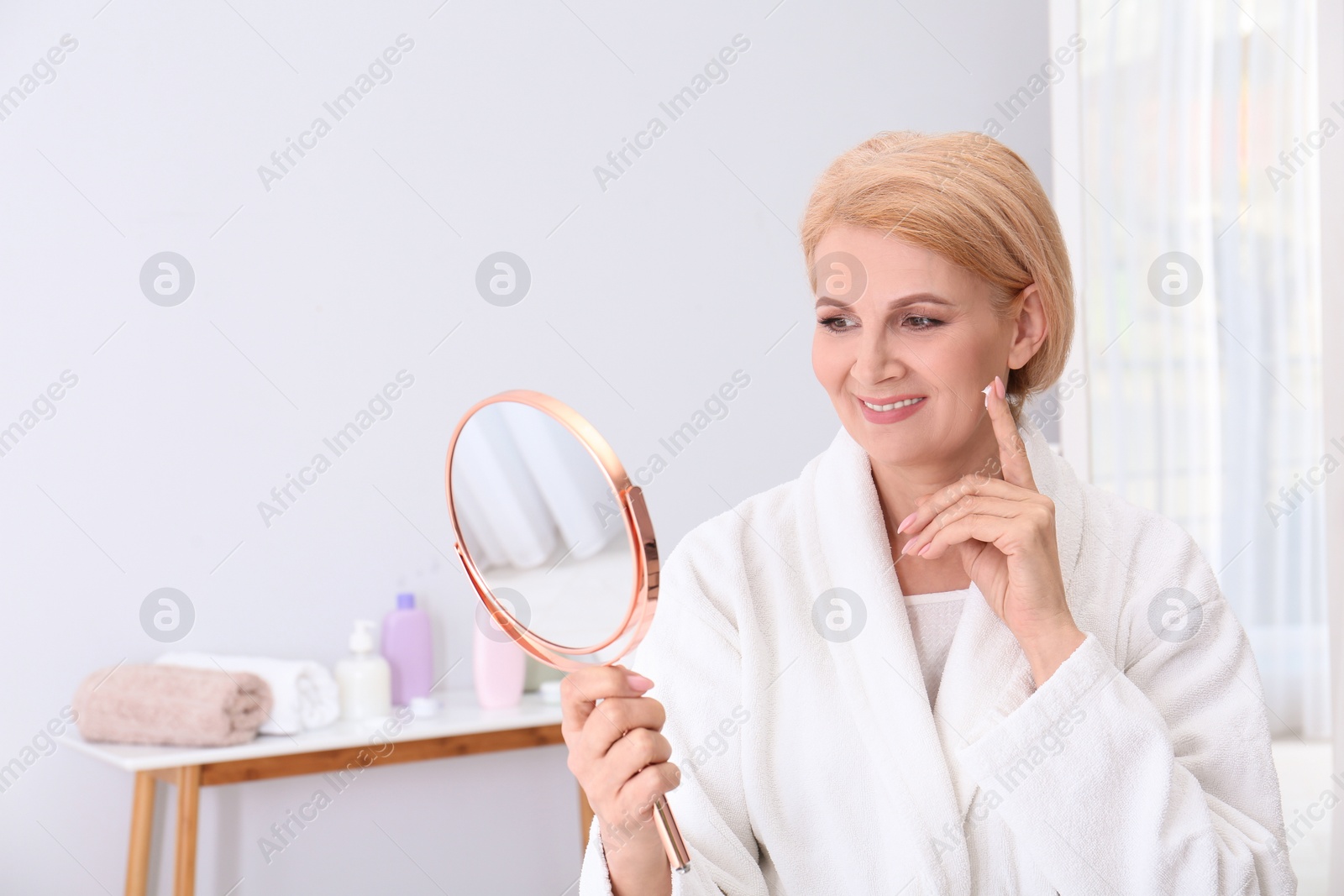 Photo of Mature woman with mirror applying face cream at home. Space for text