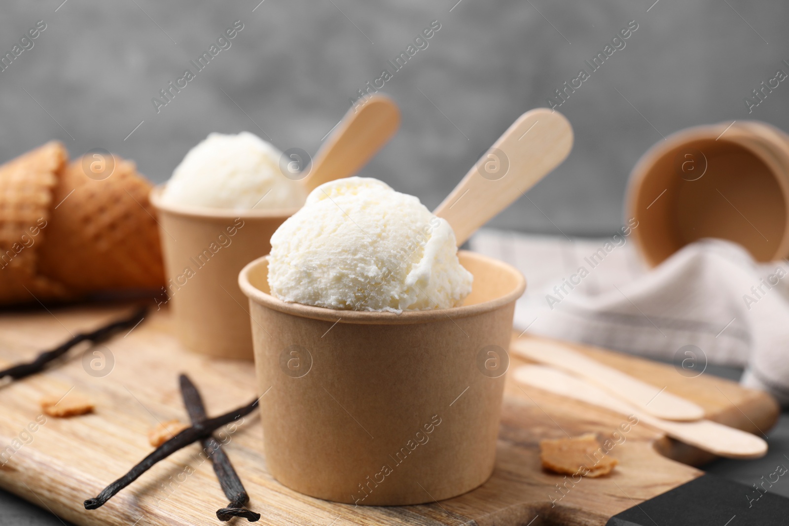 Photo of Paper cups with delicious ice cream and vanilla pods on wooden board, closeup