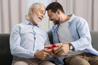 Photo of Son giving gift box to his dad on sofa at home