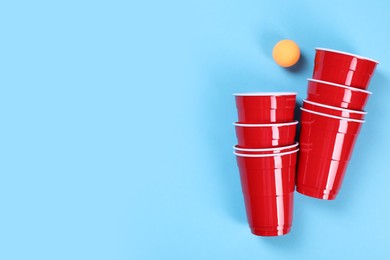 Plastic cups and ball on light blue background, flat lay with space for text. Beer pong game