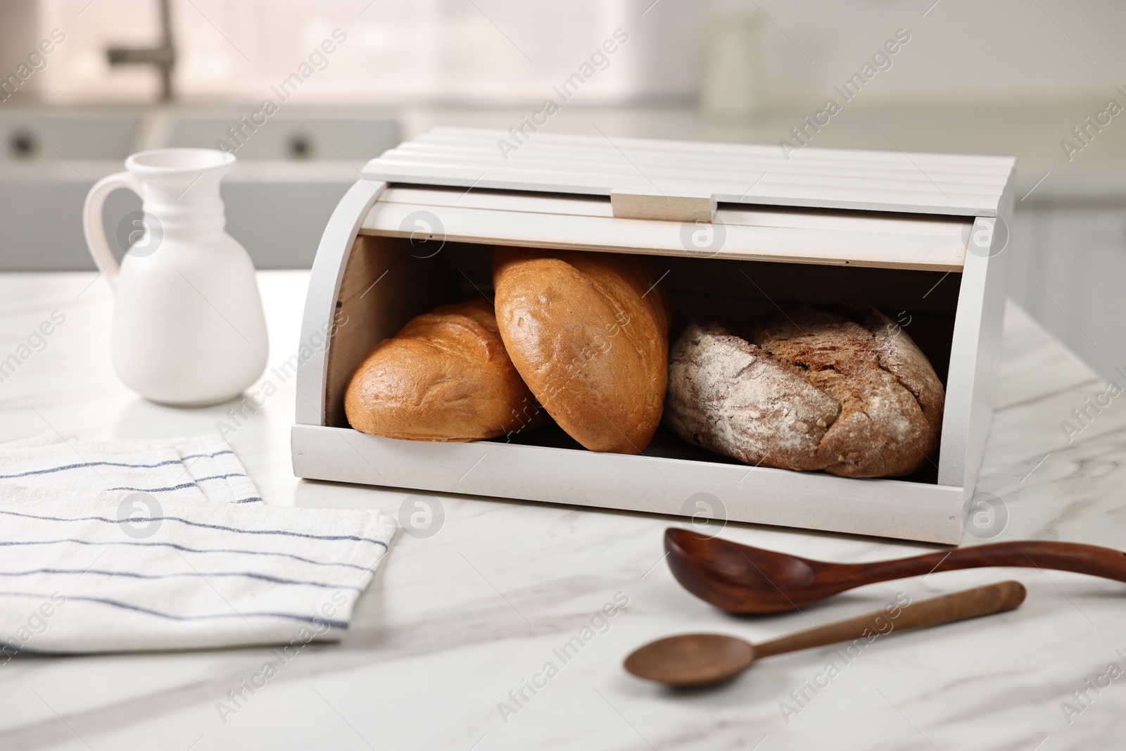 Photo of Wooden bread basket with freshly baked loaves and spoons on white marble table in kitchen