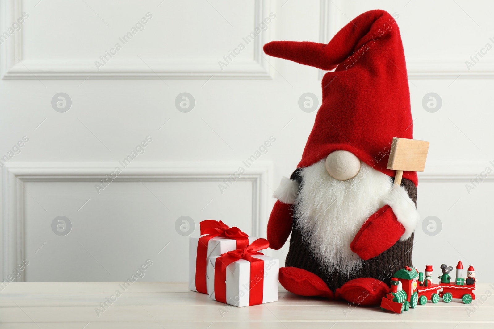 Photo of Cute Christmas gnome, gift boxes and toy train on white wooden table. Space for text