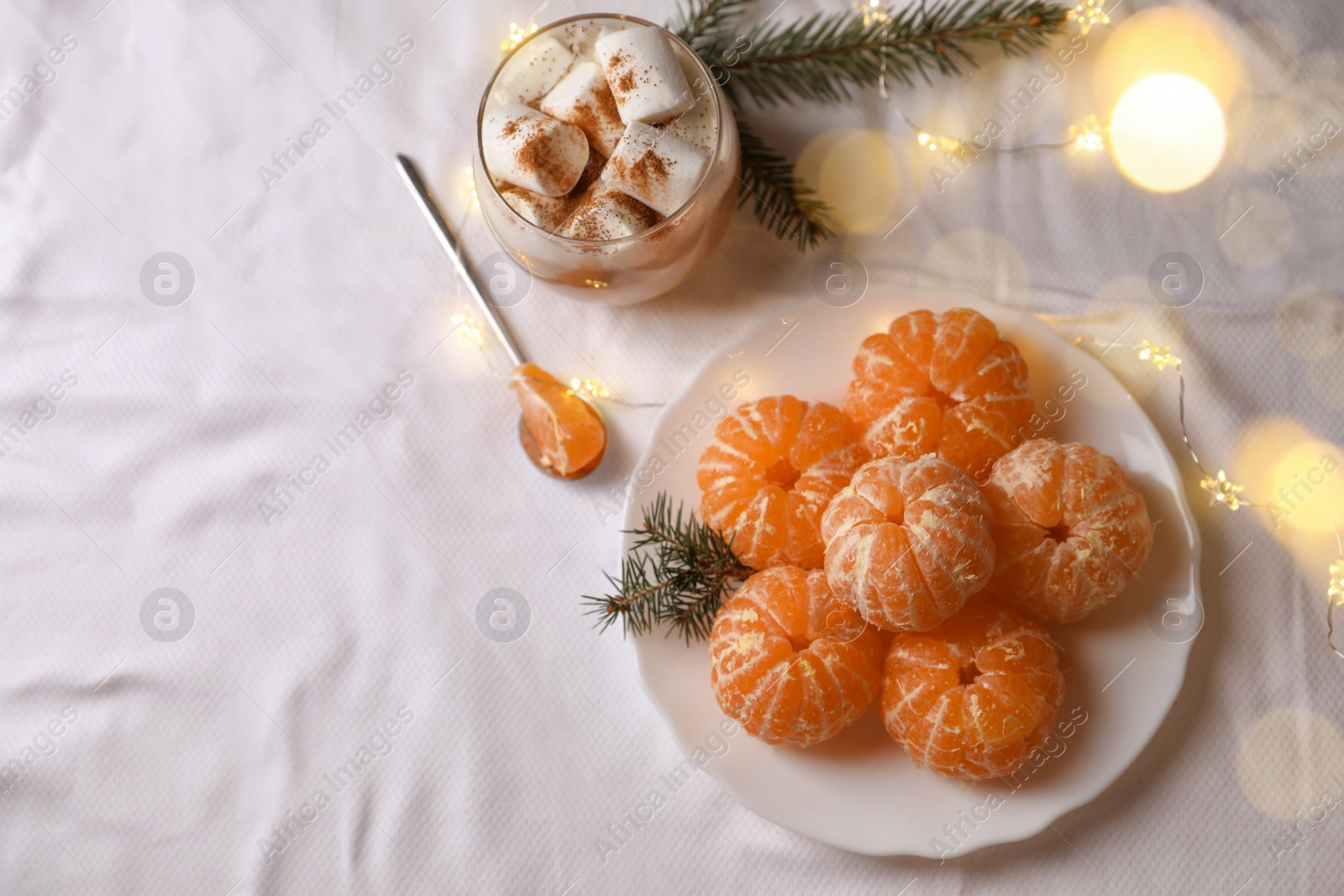 Photo of Peeled delicious ripe tangerines, festive lights and glass of drink with marshmallows on white bedsheet, flat lay. Space for text