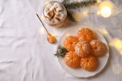 Peeled delicious ripe tangerines, festive lights and glass of drink with marshmallows on white bedsheet, flat lay. Space for text