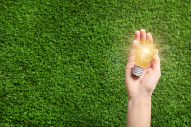 Image of Woman holding glowing light bulb over green grass, top view. Space for text