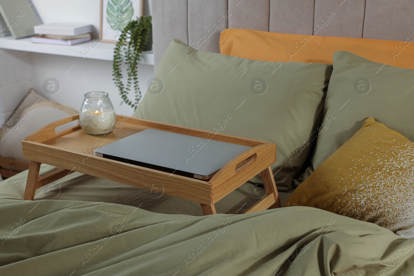 Photo of Wooden tray with modern laptop and burning candle on bed indoors