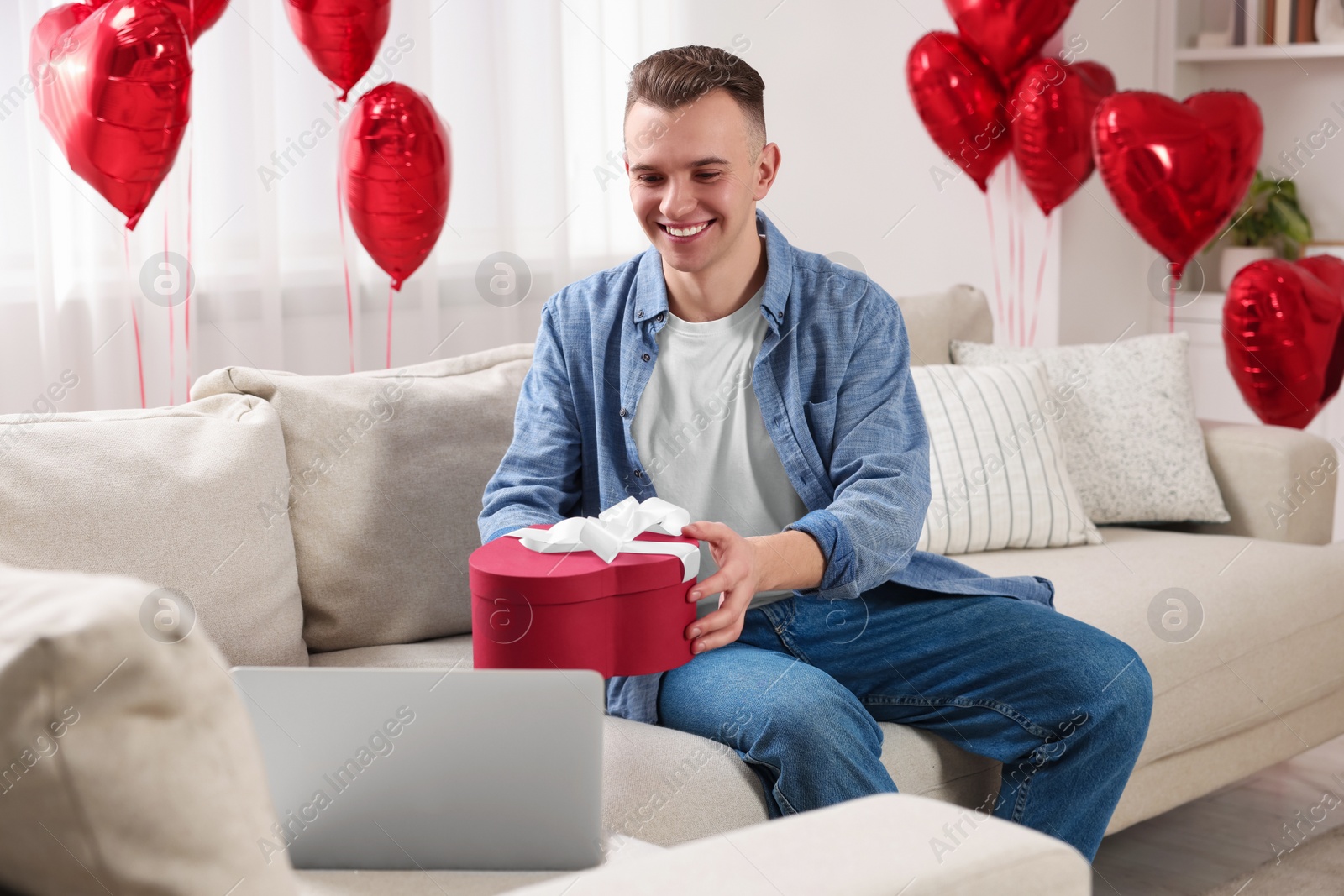 Photo of Valentine's day celebration in long distance relationship. Man holding gift box while having video chat with his girlfriend via laptop at home