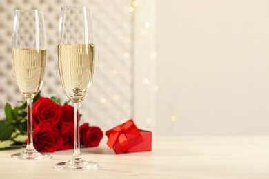 Beautiful engagement ring with gemstone in glass of sparkling wine and roses on white wooden table. Space for text