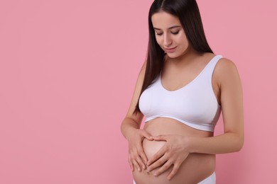 Beautiful pregnant woman in stylish comfortable underwear making heart with hands on her belly against pink background, space for text